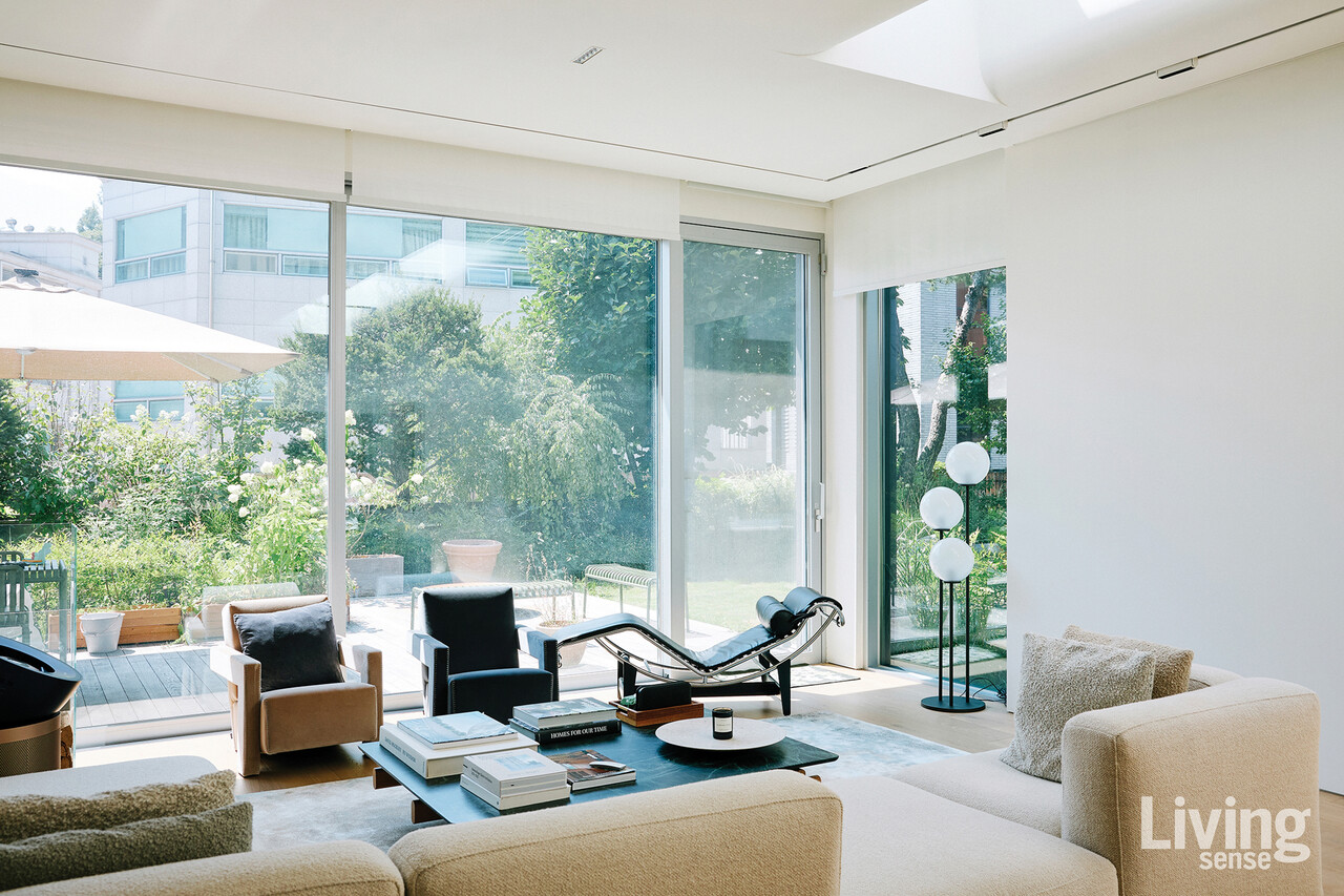 큰 창을 통해 정원의 초록과 빛이 스며드는 거실. 르 코르뷔지에의 데이베드와 미니멀한 가구들이 놓인 공간은 휴식과 풍경을 함께 즐기는 장면을 완성한다.